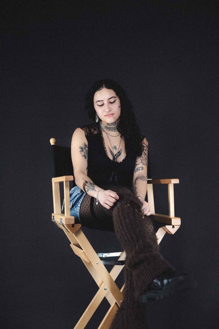 Portrait von Talia, PIK ARTIST bei PIKASS TATTOO, sitzend auf einem Regiestuhl, mit sichtbaren Tattoos an Armen und modernem, ausdrucksstarkem Look.