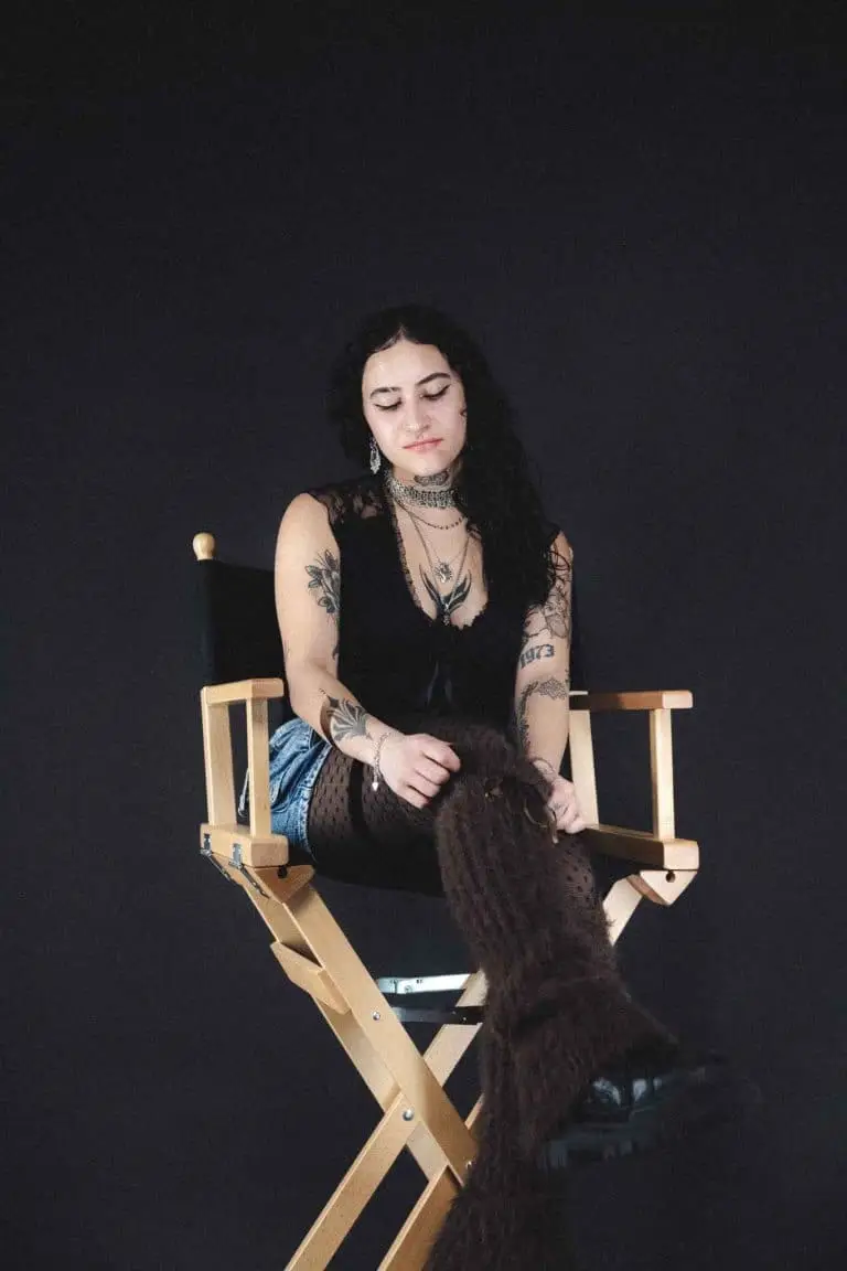 Portrait von Talia, PIK ARTIST bei PIKASS TATTOO, sitzend auf einem Regiestuhl, mit sichtbaren Tattoos an Armen und modernem, ausdrucksstarkem Look.