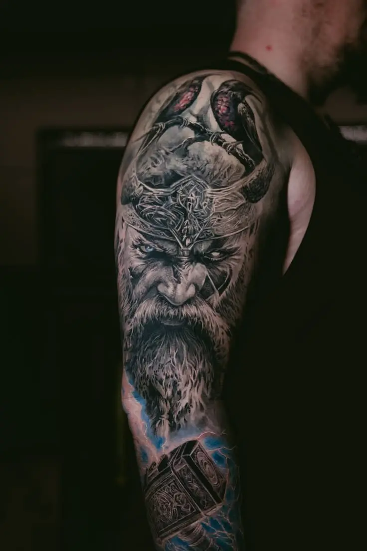 Abgeheiltes Color Realism Tattoo am Oberarm mit Wikinger-Portrait, Raben, Thorhammer und blauen Blitzeffekten