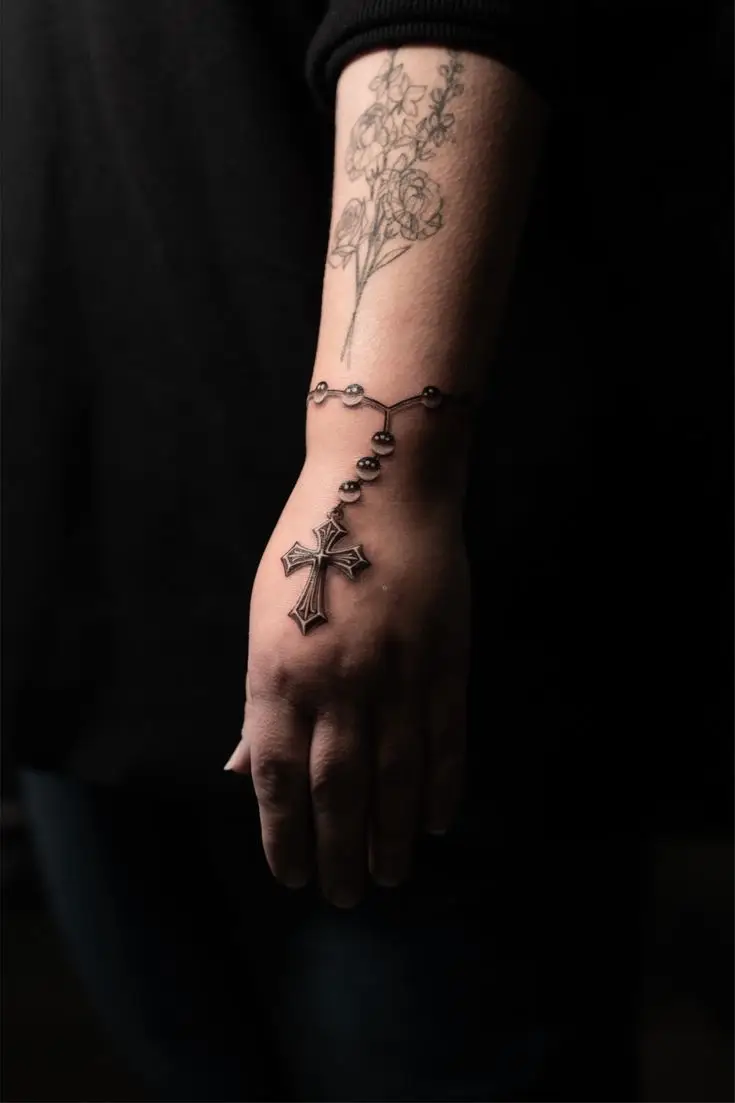 Feines Tattoo am Unterarm und Handgelenk mit Blumenmotiv und realistischem Rosenkranz mit Kreuz