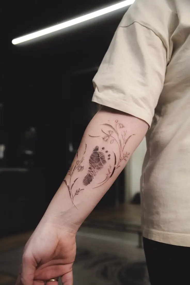 Feines Fineline Tattoo auf dem Unterarm mit kleinem Fußabdruck, Blumen und zarten floralen Linien