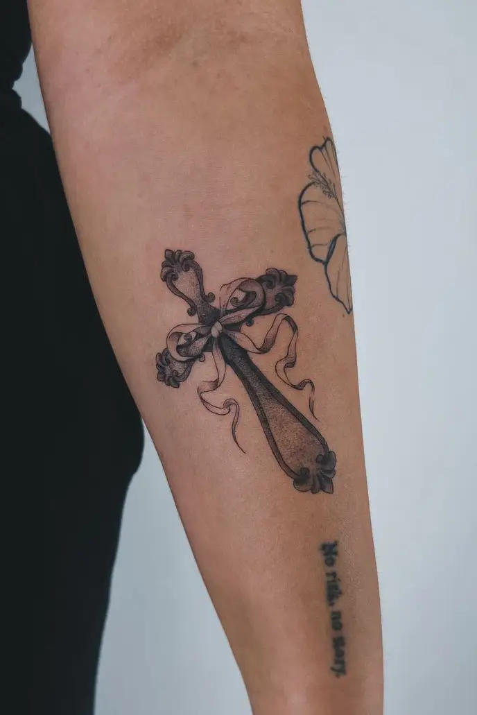 Ornamentales Kreuz-Tattoo mit Schleife und feinen Black-and-Grey-Details am Unterarm